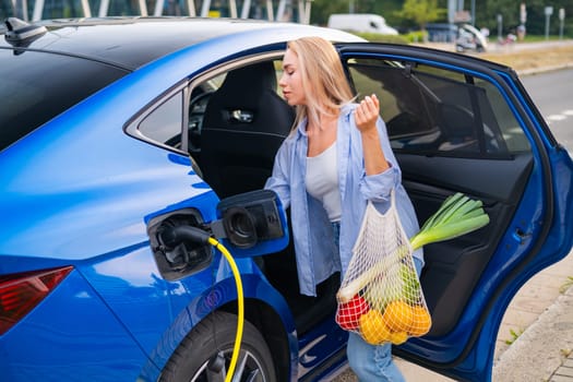 Woman charging her electric car at a public station while carrying groceries in a bag from the shop