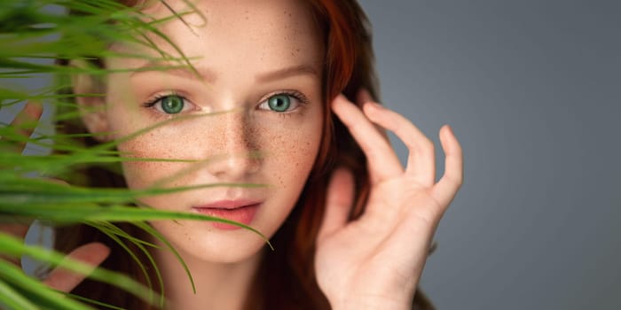 Natural Haircare. Ginger-Haired Young Woman Touching Red Hair Posing With Green Plant Leaves Near Face On Gray Background In Studio. Beauty And Facial Skin Care Cosmetics. Selective Focus