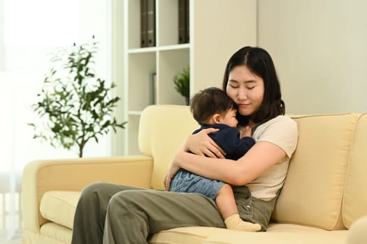 Loving young mother cuddling her baby boy sitting on sofa. Parenting, childcare and family concept.