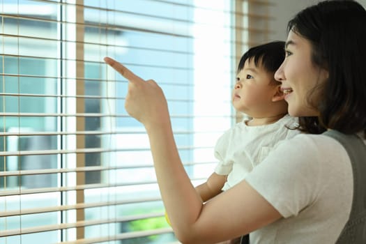 Smiling mother holding curiosity baby boy and pointing out something outside the window.