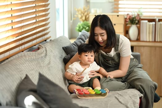 Beautiful young mother and little baby boy playing with toys on sofa at home. Parenting, childcare and family concept..