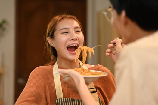 Young man feeding his girlfriend with tasty spaghetti in kitchen.