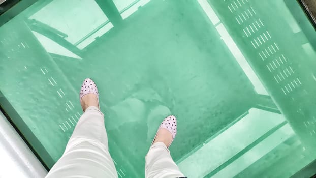 Feet rest on a transparent glass floor of a cable car, revealing vibrant greenery below in a stunning aerial view