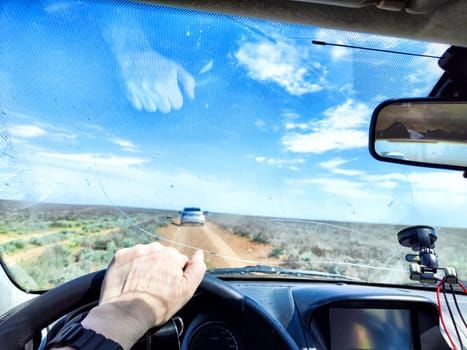 A driver navigates an open road with a cracked windshield while surrounded by green fields and a bright blue sky filled with clouds.
