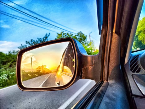 A vibrant sunset is visible in the side mirror while driving on a quiet country road.