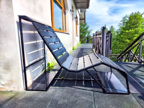 A sleek lounge chair made of black slats rests on a terrace, enhanced by natural light and lush green surroundings.