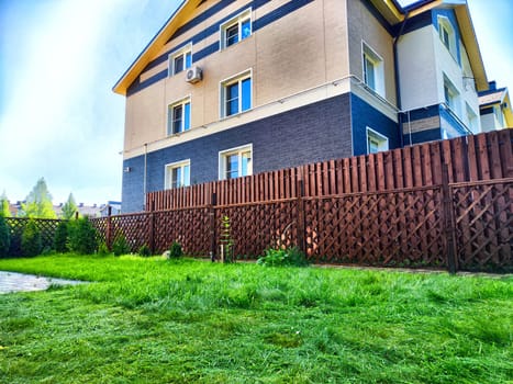 A well-maintained green lawn with a wooden fence stands next to a modern residential building under blue skies.
