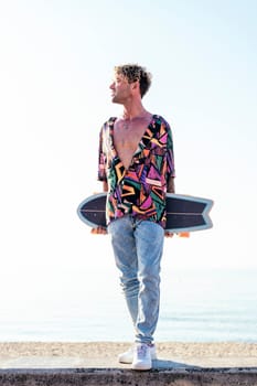 young hipster male skater in trendy summer outfit carrying skateboard while standing on embankment against sea and cloudless sky in summer day