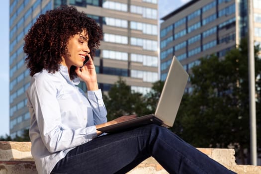 side view of focused happy female entrepreneur sitting on stone bench in park and working remotely while speaking on smartphone and using laptop