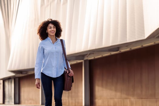 satisfied ethnic businesswoman with bag walking along street and smiling while looking forward