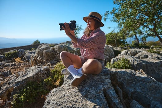 A woman in casual attire is sitting on rocks, capturing the scenic landscape with a camera under a clear blue sky.