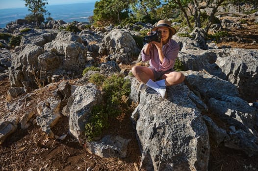 A woman sits on rocky terrain, capturing moments with a camera, embracing nature's beauty under sunlight.