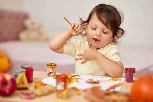 smiling little child girl painting her hands and autumn leaves. baby girl drawing hands in yellow paint