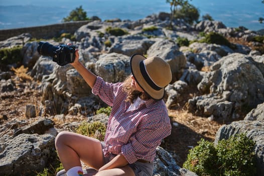 A woman in a straw hat sits among rocks taking a photo with a DSLR camera. She is enjoying the scenic mountain view under the sunlight.