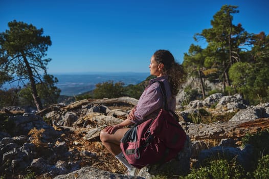 A woman sits on a rocky terrain, absorbing the scenic beauty of a mountainous landscape. Surrounded by trees, she carries a backpack, embodying tranquility and adventure.
