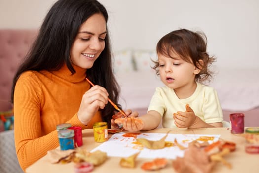 Cute little girl and mom spending good time painting hands and autumn yellow leaves together
