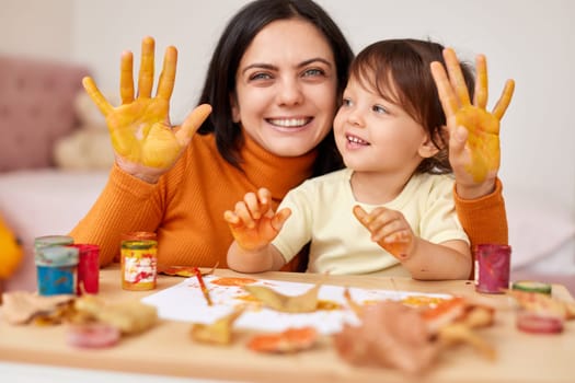 smiling little child girl and mother spending good time painting hands and autumn leaves together. mom and baby showing hands in yellow paint