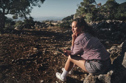 A woman sits on a rock in a scenic outdoor setting, enjoying the peaceful nature. She holds a smartphone and seems deep in thought or contemplation, under a clear blue sky.