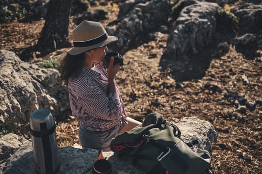 A young woman in a hat sips coffee while sitting on a rock outdoors, surrounded by nature. She is dressed casually with a backpack and thermos nearby, enjoying a peaceful hiking moment.