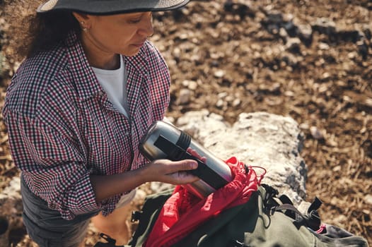 A woman in casual wear, taking moment to enjoy a hot drink from thermos during a peaceful outdoor hike. She is surrounded by natural scenery and carrying a backpack, highlighting leisure and adventure