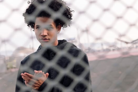 portrait of thoughtful teen african american male with curly hair standing behind metal chain link fence on urban street, concept of adolescence and mental health