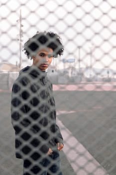 thoughtful teen african american male with curly hair looking at camera while standing behind metal chain link fence on urban street, concept of adolescence and mental health
