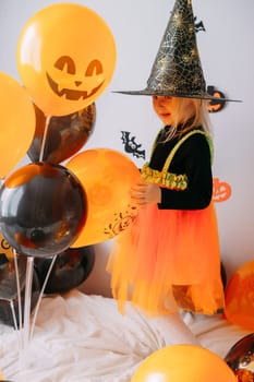 Children's Halloween - a girl in a witch hat and a carnival costume with airy orange and black balloons at home. Ready to celebrate Halloween.