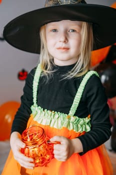Children's Halloween - a girl in a witch hat and a carnival costume with airy orange and black balloons at home. Ready to celebrate Halloween.