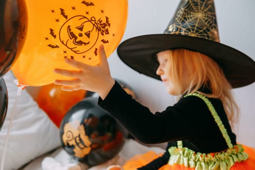 Children's Halloween - a girl in a witch hat and a carnival costume with airy orange and black balloons at home. Ready to celebrate Halloween.