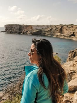 A woman in sunglasses overlooking a rocky coastline on a sunny day, latin woman looking out over the mediterranean sea cliffs, in mallorca balearic islands spain,