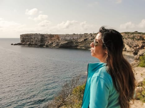 Woman with long hair gazes at ocean from a rocky cliff under sunny skies, latin woman looking out over the mediterranean sea cliffs, in mallorca balearic islands spain,