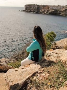 Woman sitting on rocky cliff overlooking the ocean on a sunny day, latin woman looking out over the mediterranean sea cliffs, in mallorca balearic islands spain,
