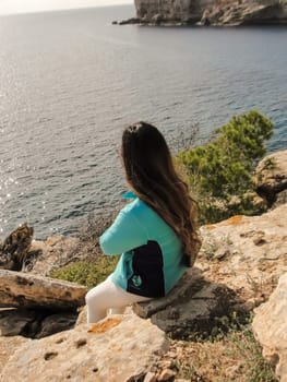 Woman sitting on rocks admiring the ocean view from a cliff, latin woman looking out over the mediterranean sea cliffs, in mallorca balearic islands spain,