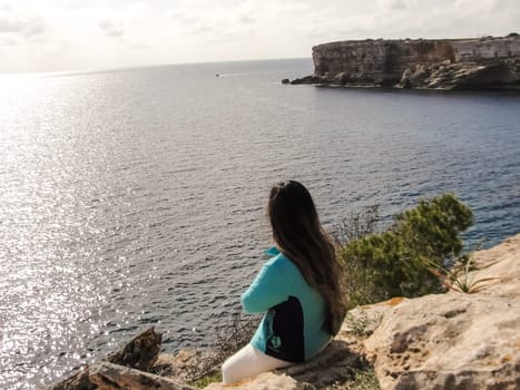 Woman perched on a cliff edge, viewing the expansive ocean under the sun, latin woman looking out over the mediterranean sea cliffs, in mallorca balearic islands spain,