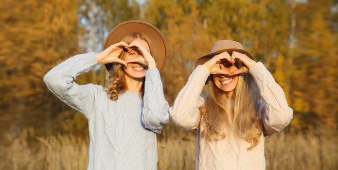 Portrait of happy young two women friends showing heart shaped sign gesture with hands, smiling girlfriends together outdoors in park, sunny day, fall season
