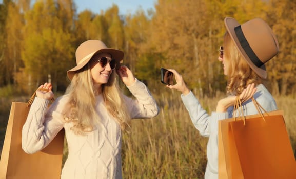 Happy two women friends taking selfie with phone in autumn park, girlfriends smiling holding shopping bags together outdoors in sunny warm fall season