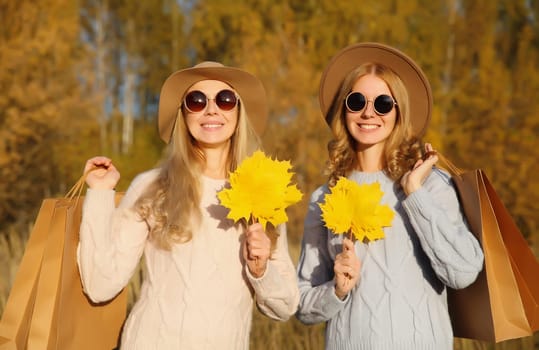 Happy young two women friends with autumn yellow leaves, holding shopping bags, stylish smiling girlfriends standing together outdoors, in sunny park, warm fall season