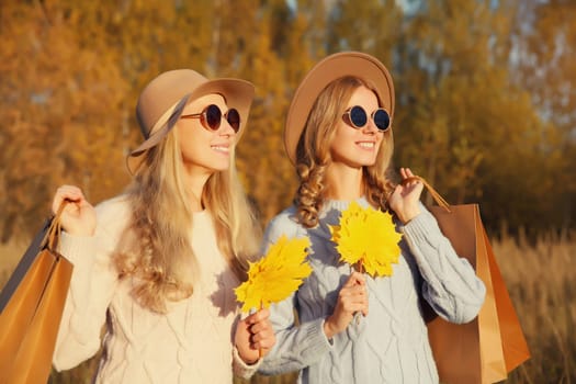 Happy young two women friends with autumn yellow leaves, holding shopping bags, stylish smiling girlfriends standing together outdoors, in sunny park, warm fall season