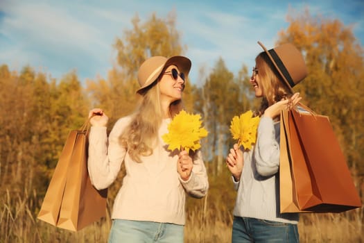 Happy young two women friends with autumn yellow leaves, holding shopping bags, stylish smiling girlfriends standing together outdoors, in sunny park, warm fall season
