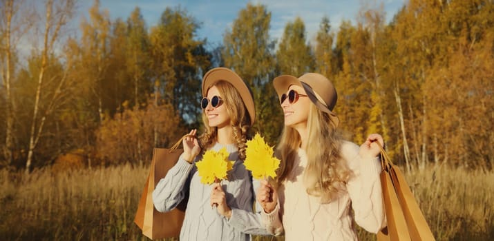 Happy young two women friends with autumn yellow leaves, holding shopping bags, stylish smiling girlfriends standing together outdoors, in sunny park, warm fall season