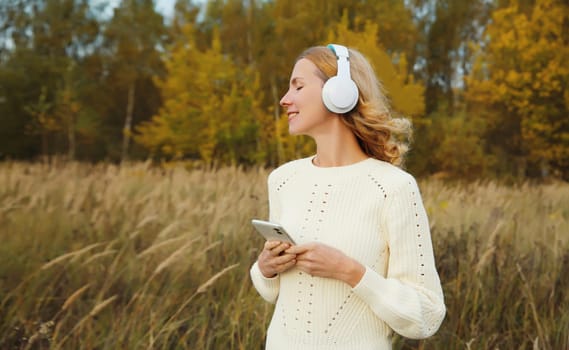 Happy inspired young woman enjoying listening to music in headphones with phone outdoors in autumn forest with yellow leaves