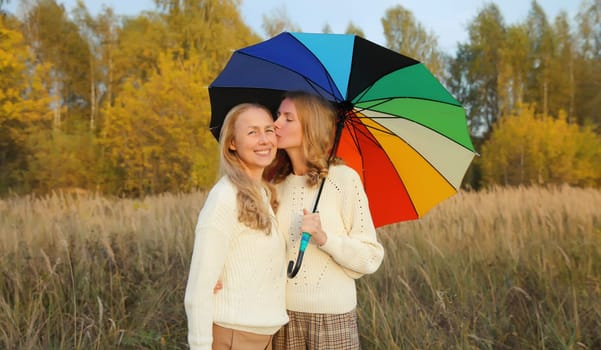 Adult daughter kissing her happy smiling mature mother holding colorful umbrella walking in autumn park with yellow leaves