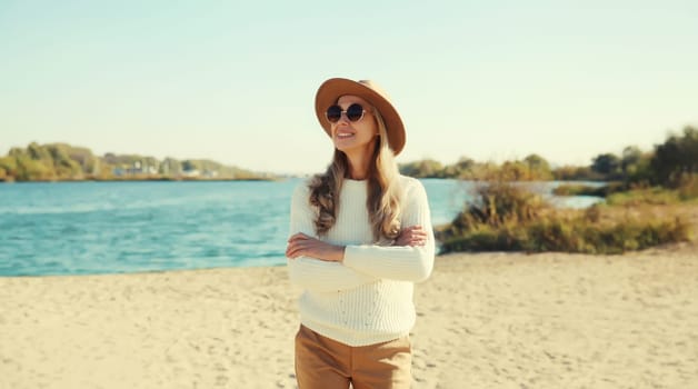 Stylish mature woman on coast beach, fashionable middle aged female model posing outdoors, wearing sweater, hat and glasses, standing with crossed arms looking away