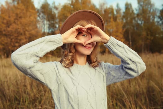 Autumn beauty portrait of stylish happy young woman showing heart shaped sign gesture with hands, fashionable girl smiling in hat, sweater outdoors in park, warm fall season