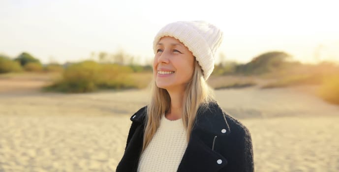 Happy stylish mature woman on coast beach, middle aged female smiling standing outdoors, wearing hat, looking away