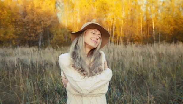 Happy smiling mature woman hugs herself in autumn park enjoying warm weather on sunny day outdoors.