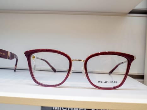 Stylish michael kors eyeglasses with dark red frames are displayed on a white shelf in an eyewear store, ready for customers to try on
