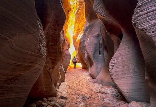 A female hiker walking through the narrow Buckskin Gulch slot canyon on the Arizona, Utah border. She hikes toward the light at the end of the canyon with the tall canyon walls glowing orange - USA