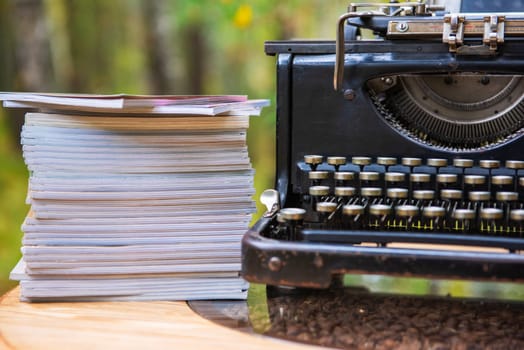 Close-up of an old-fashioned typewriter beside a tall stack of papers, set outdoors with a blurred natural background.