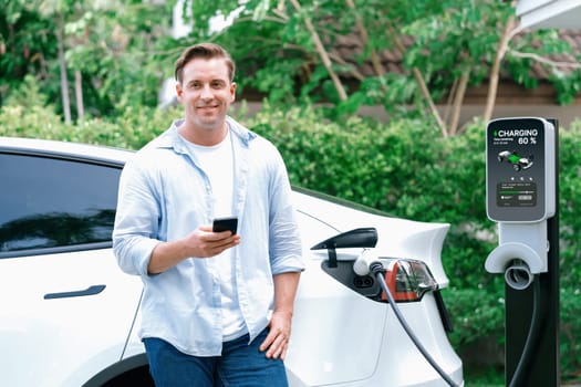 Modern eco-friendly man recharging electric vehicle from home EV charging station. Innovative EV technology utilization for tracking energy usage to optimize battery charging at home. Synchronos